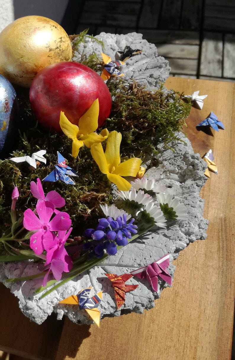 May include: A concrete nest with moss, decorated with origami birds, flowers, and painted Easter eggs. The nest is on a wooden surface.