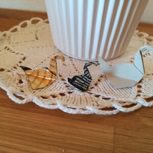May include: Two origami swans made from paper wrappers. One swan is gold and the other is black and white. The swans are sitting on a white crocheted doily.