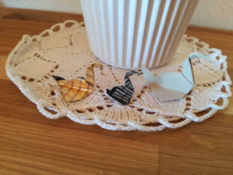 May include: Two origami swans made from paper wrappers. One swan is gold and the other is black and white. The swans are sitting on a white crocheted doily.