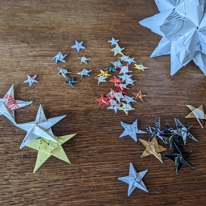 May include: A collection of origami stars in various sizes and colors, arranged on a wooden surface. The stars are made from paper, some with printed text, and range from white to yellow, red, and black. A large, multi-pointed star is also present.