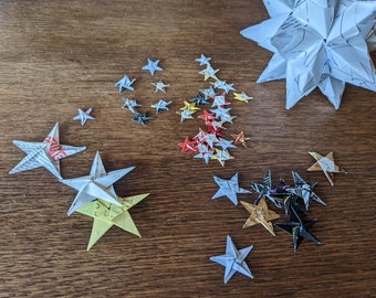 May include: A collection of origami stars in various sizes and colors, arranged on a wooden surface. The stars are made from paper, some with printed text, and range from white to yellow, red, and black. A large, multi-pointed star is also present.
