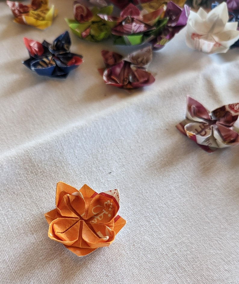 May include: A close-up of an orange origami flower with a red and white pattern. The flower is made from paper and has five petals.