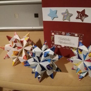 Puede incluir: Tres estrellas de origami hechas de papel colorido, con una tarjeta roja en el fondo. La tarjeta tiene tres decoraciones en forma de estrella y el texto "Fr&ouml;hliche Weihnachten". Las estrellas son blancas, azules y rojas.