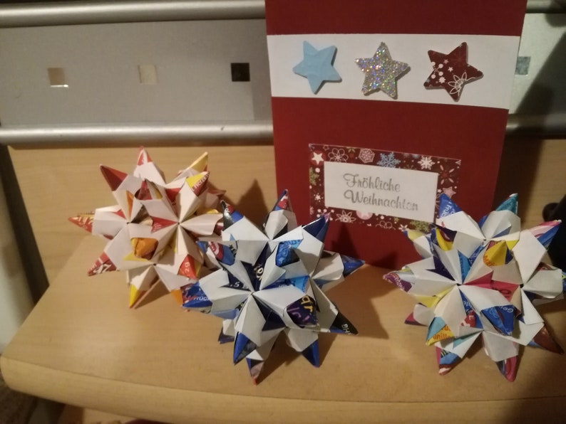 Puede incluir: Tres estrellas de origami hechas de papel colorido, con una tarjeta roja en el fondo. La tarjeta tiene tres decoraciones en forma de estrella y el texto "Fr&ouml;hliche Weihnachten". Las estrellas son blancas, azules y rojas.