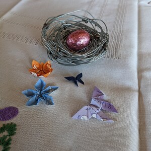 May include: A wire nest with a pink chocolate egg, surrounded by origami butterflies and flowers in blue, orange, and purple.