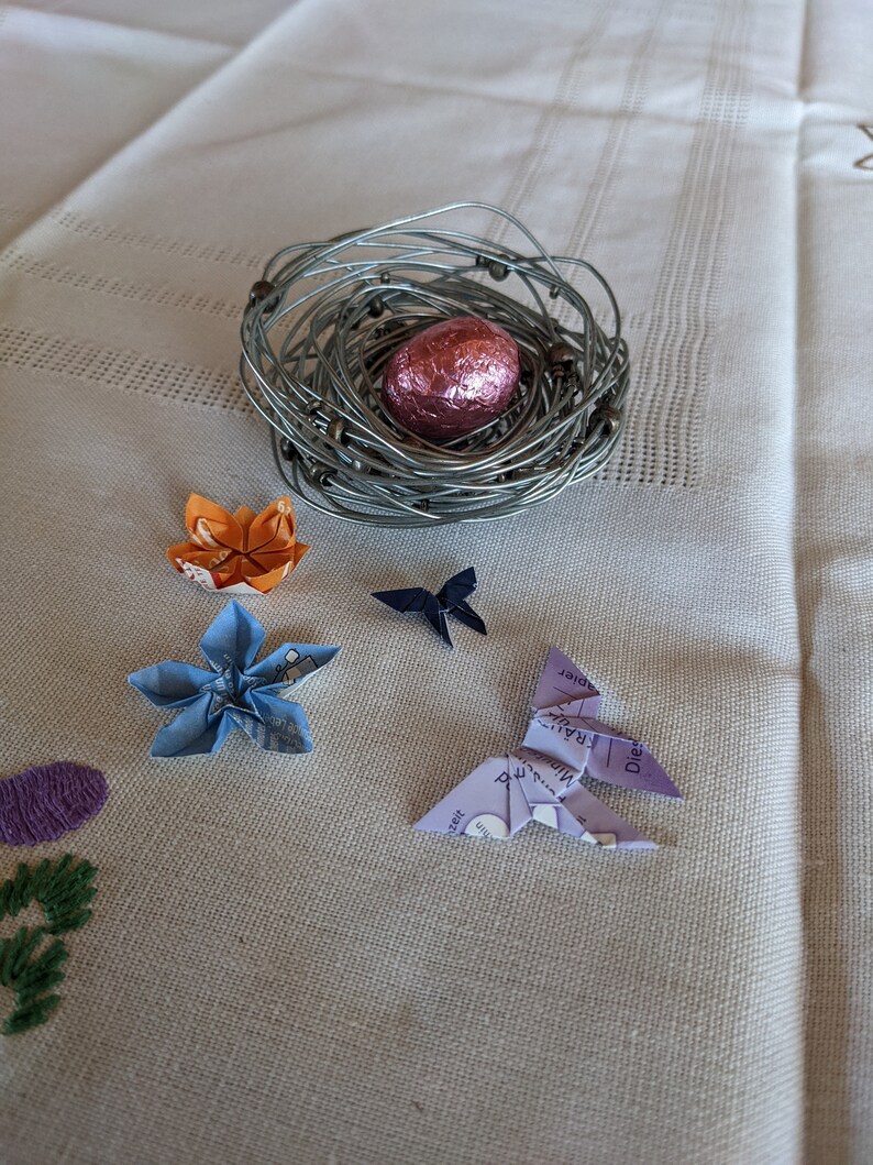 May include: A wire nest with a pink chocolate egg, surrounded by origami butterflies and flowers in blue, orange, and purple.