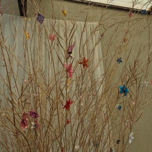 May include: A collection of colorful origami paper flowers are displayed on a dried branch arrangement. The flowers are in shades of blue, pink, red, yellow, and green.