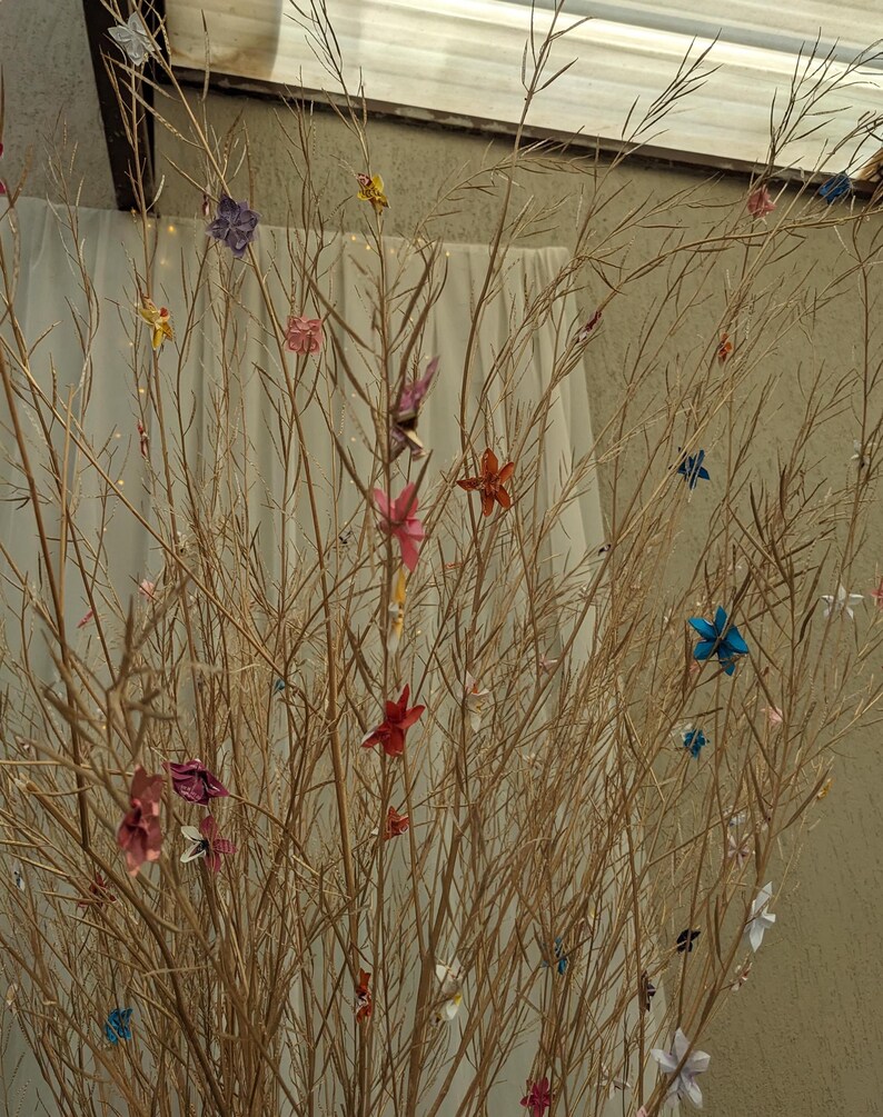 May include: A collection of colorful origami paper flowers are displayed on a dried branch arrangement. The flowers are in shades of blue, pink, red, yellow, and green.