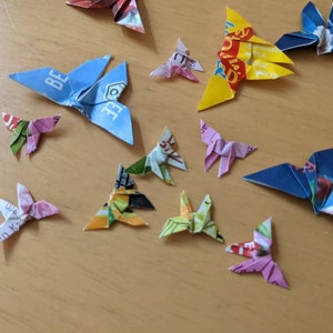May include: A collection of origami butterflies made from colorful paper. The butterflies are in various colors, including blue, yellow, pink, and green. Some of the butterflies have text on them, such as "Be" and "Wig".