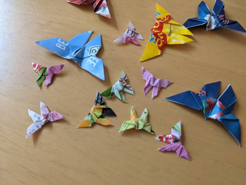 May include: A collection of origami butterflies made from colorful paper. The butterflies are in various colors, including blue, yellow, pink, and green. Some of the butterflies have text on them, such as "Be" and "Wig".