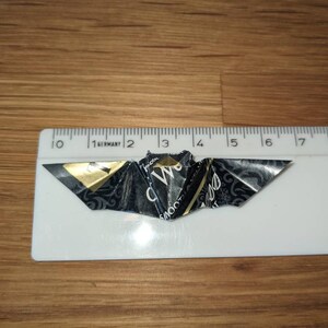 Puede incluir: Un murci&eacute;lago de origami hecho a mano, elaborado con papel negro y dorado, descansa sobre una superficie de madera junto a una regla. La envergadura del murci&eacute;lago es de aproximadamente 5 cm.
