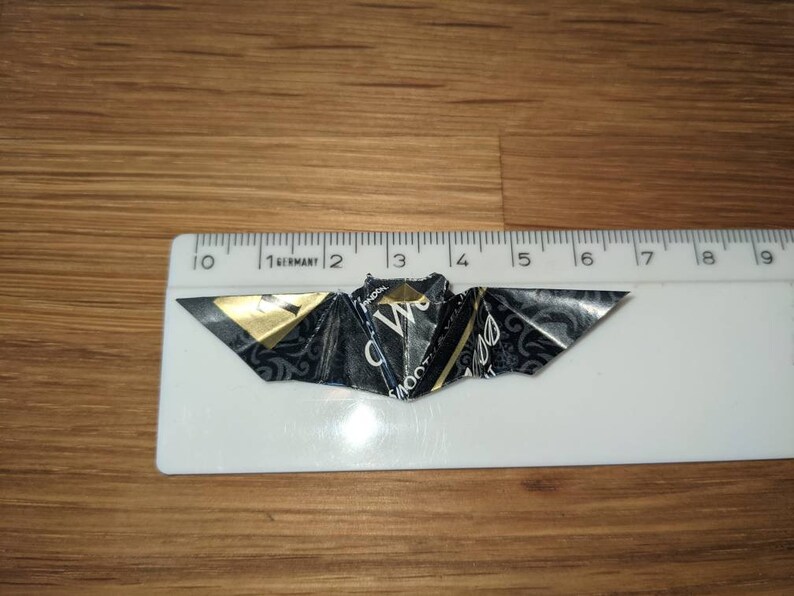 Puede incluir: Un murci&eacute;lago de origami hecho a mano, elaborado con papel negro y dorado, descansa sobre una superficie de madera junto a una regla. La envergadura del murci&eacute;lago es de aproximadamente 5 cm.