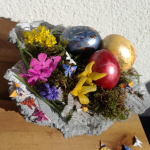 May include: A gray concrete bowl filled with moss, flowers, and colorful Easter eggs.  The bowl is decorated with origami butterflies.