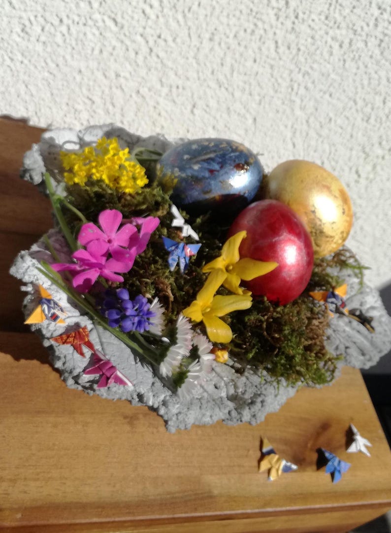 May include: A gray concrete bowl filled with moss, flowers, and colorful Easter eggs.  The bowl is decorated with origami butterflies.