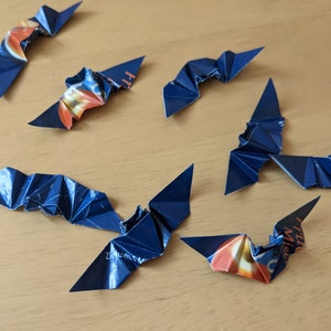 Puede incluir: Murci&eacute;lagos de origami hechos a mano con papel azul oscuro y naranja. Los murci&eacute;lagos est&aacute;n dispuestos sobre una superficie de madera de color marr&oacute;n claro. Las alas est&aacute;n dobladas para crear un efecto texturizado, y los murci&eacute;lagos est&aacute;n en vuelo.