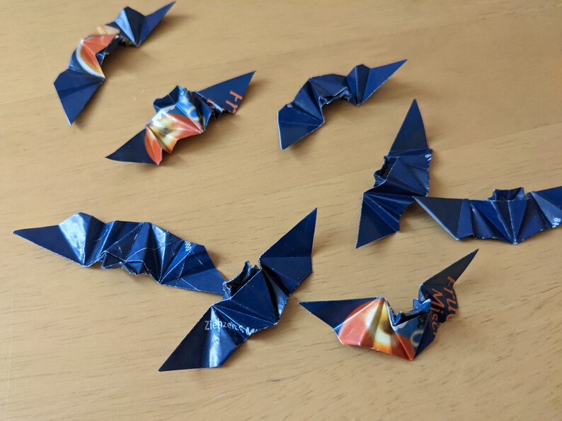 Puede incluir: Murci&eacute;lagos de origami hechos a mano con papel azul oscuro y naranja. Los murci&eacute;lagos est&aacute;n dispuestos sobre una superficie de madera de color marr&oacute;n claro. Las alas est&aacute;n dobladas para crear un efecto texturizado, y los murci&eacute;lagos est&aacute;n en vuelo.