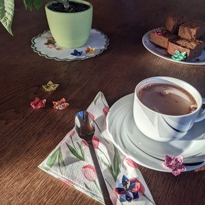 May include: A white cup with a saucer filled with hot chocolate, a spoon, and a napkin with a floral pattern. There are origami paper flowers on the table, a potted plant, and a plate of chocolate brownies.