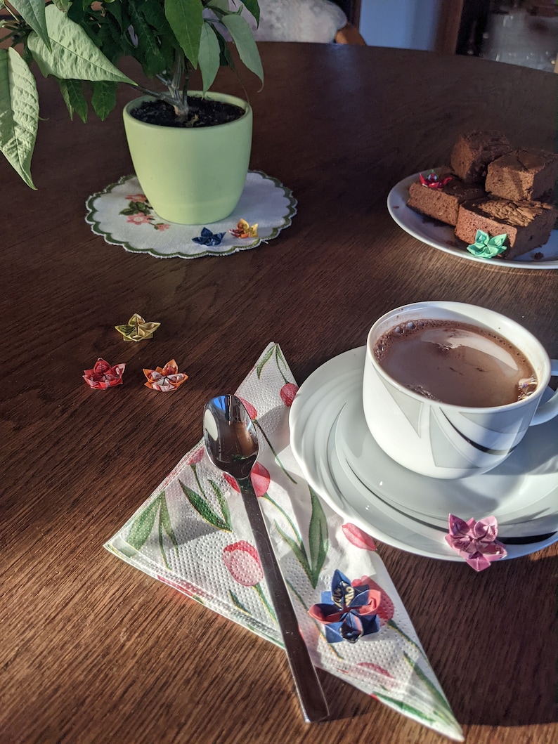 May include: A white cup with a saucer filled with hot chocolate, a spoon, and a napkin with a floral pattern. There are origami paper flowers on the table, a potted plant, and a plate of chocolate brownies.