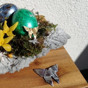 May include: A green Easter egg with a gold origami butterfly on top, surrounded by moss and yellow flowers, sits in a gray concrete nest. A silver origami butterfly is on a wooden surface.