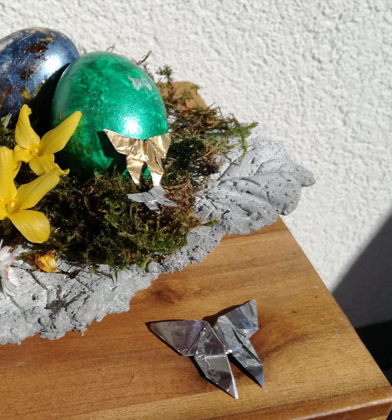 May include: A green Easter egg with a gold origami butterfly on top, surrounded by moss and yellow flowers, sits in a gray concrete nest. A silver origami butterfly is on a wooden surface.