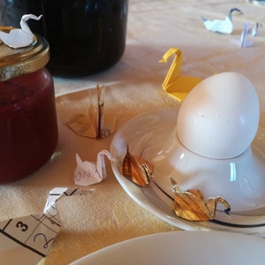 May include: A white egg in an egg cup on a white saucer with gold origami swans on a white tablecloth. There are other origami swans on the table, some are white and some are yellow.