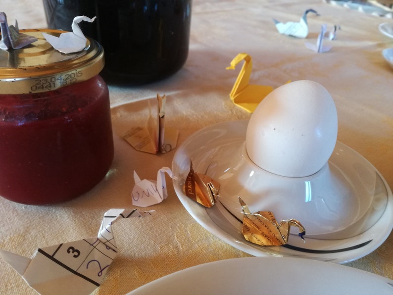 May include: A white egg in an egg cup on a white saucer with gold origami swans on a white tablecloth. There are other origami swans on the table, some are white and some are yellow.