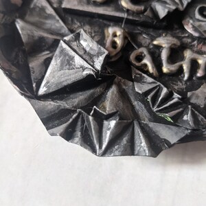 Puede incluir: Una obra de arte met&aacute;lica texturizada, de color gris oscuro, con letras en relieve de color dorado. La obra de arte tiene una apariencia plegada, como un origami. El texto es parcialmente visible, con las letras "BE" y "ATH" discernibles.