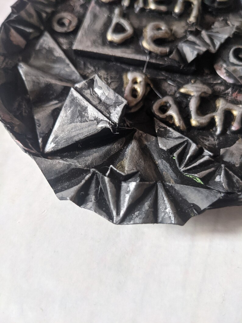Puede incluir: Una obra de arte met&aacute;lica texturizada, de color gris oscuro, con letras en relieve de color dorado. La obra de arte tiene una apariencia plegada, como un origami. El texto es parcialmente visible, con las letras "BE" y "ATH" discernibles.