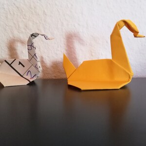 May include: Two origami swans, one white and one yellow, are sitting on a black surface. The white swan is made from a piece of paper with numbers written on it.