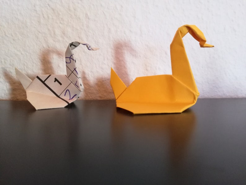 May include: Two origami swans, one white and one yellow, are sitting on a black surface. The white swan is made from a piece of paper with numbers written on it.