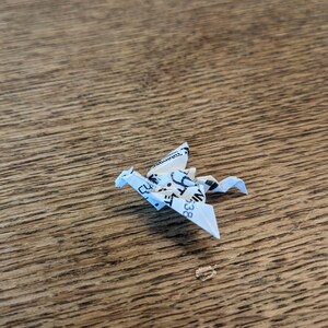Może przedstawiać: Biały origami smok złożony z kawałka papieru z czarnym tekstem. Smok siedzi na drewnianej powierzchni.