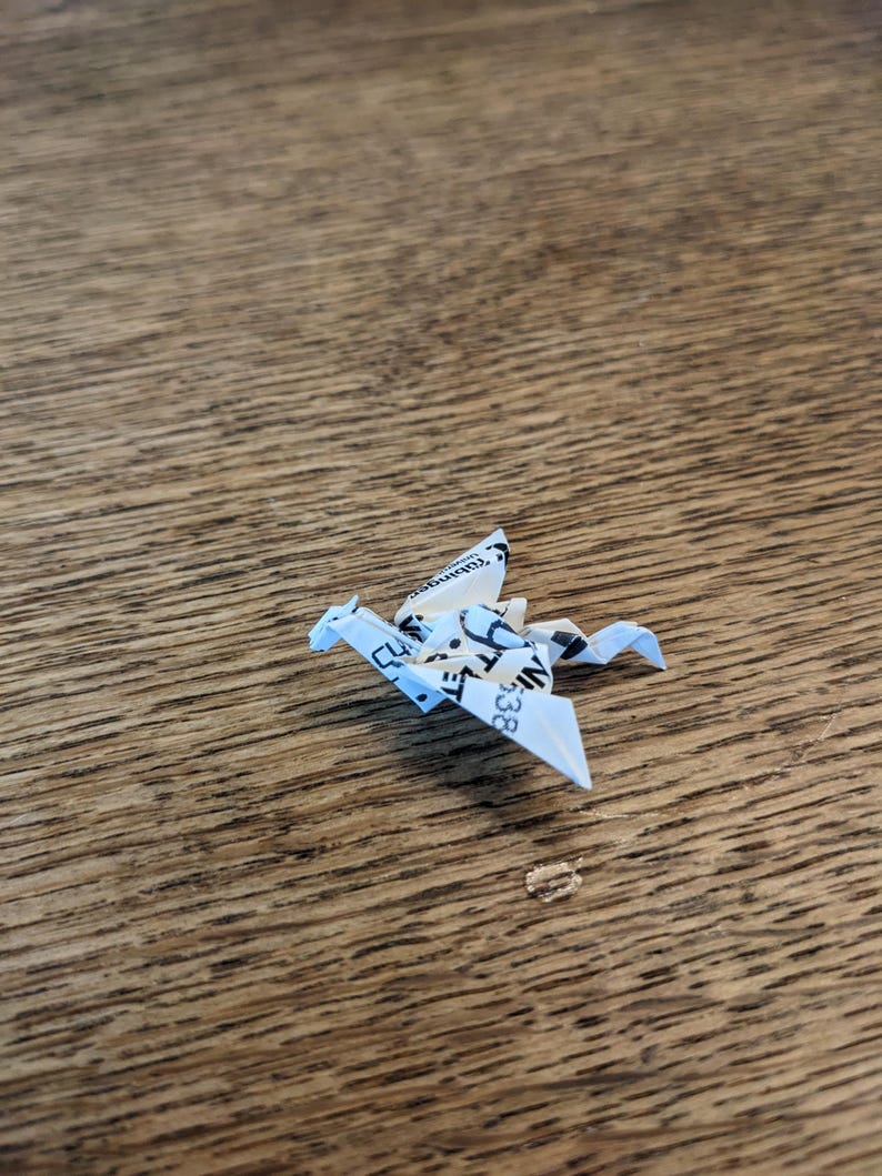 Może przedstawiać: Biały origami smok złożony z kawałka papieru z czarnym tekstem. Smok siedzi na drewnianej powierzchni.