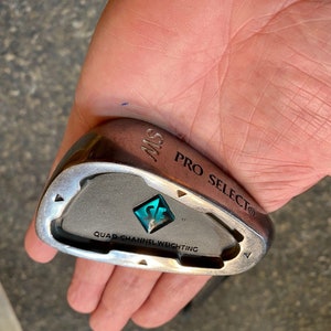 May include: A silver golf club with the text "PRO SELECT®" and "QUAD-CHANNEL WEIGHTING" on the face. The club has a teal blue logo with the letters "SE" inside a diamond shape.