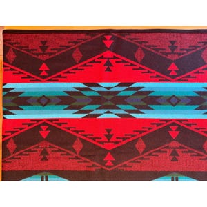 May include: A rectangular textile with a Southwestern design. The pattern features red, black, and turquoise geometric shapes, including triangles and diamonds. The textile has a border and is likely a blanket or rug.