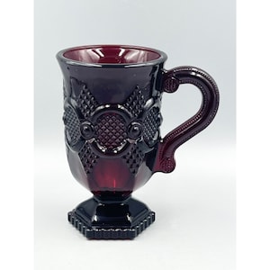 Puede incluir: Una taza de vidrio rojo oscuro con una base con pie y un asa decorativa. La taza tiene un patrón de diamante y un diseño de círculo en relieve.
