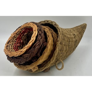 May include: A woven cornucopia basket in shades of brown, tan, and red. The basket is shaped like a horn, with a wide opening and a tapered end. The basket has a braided rim and a woven body. A ring handle is attached to the side.