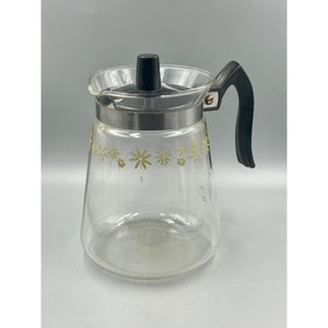 May include: A clear glass coffee pot with a black handle and lid. The pot has a silver band and a yellow floral pattern. The pot has measurement markings. The pot is a classic design.