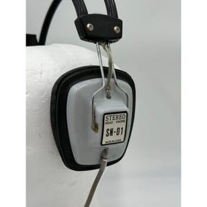Puede incluir: Auriculares est&eacute;reo vintage con una copa de oreja blanca de forma cuadrada y borde negro. Una etiqueta en el lateral dice STEREO HEAD-PHONE SN-01 MADE IN JAPAN. El dispositivo incluye una diadema negra y un marco de alambre met&aacute;lico plateado.