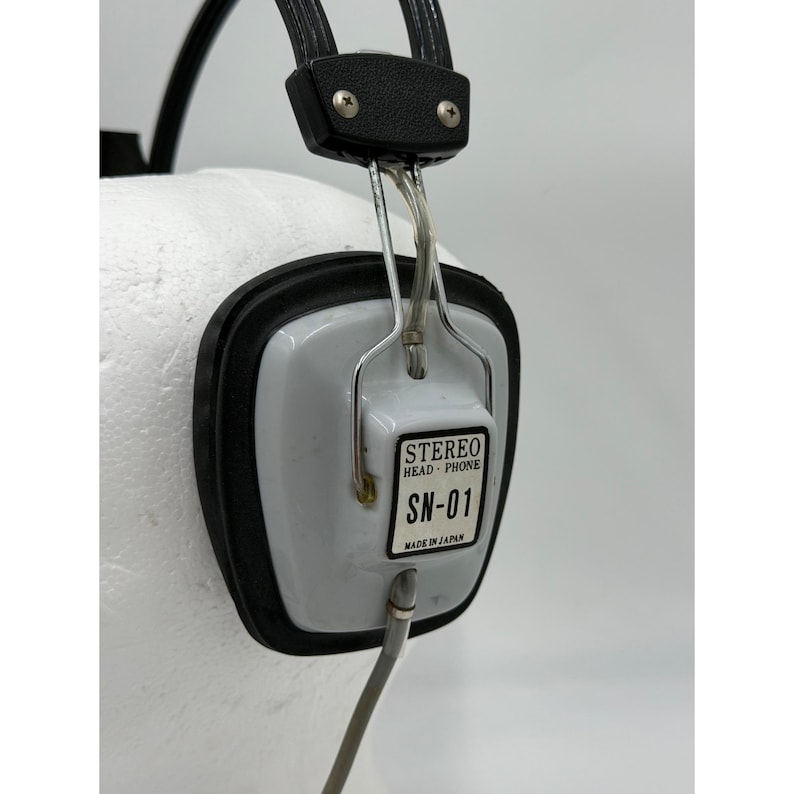 Puede incluir: Auriculares est&eacute;reo vintage con una copa de oreja blanca de forma cuadrada y borde negro. Una etiqueta en el lateral dice STEREO HEAD-PHONE SN-01 MADE IN JAPAN. El dispositivo incluye una diadema negra y un marco de alambre met&aacute;lico plateado.