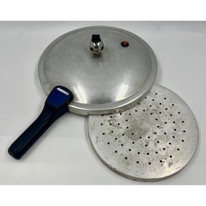 Puede incluir: Una olla a presión Mirro de color plateado con un asa negra y un inserto de metal perforado. La tapa tiene una válvula de presión y un indicador rojo. El logotipo de Mirro es visible.