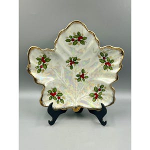 May include: A white ceramic leaf-shaped serving dish with a pearlescent glaze and gold trim. The dish is decorated with green holly leaves and red berries.