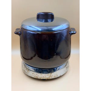 May include: A vintage brown ceramic slow cooker with a silver lid and base. The lid has a dark brown handle. The slow cooker has two side handles and is sitting on a silver base with some wear.