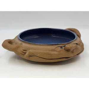 May include: A handmade ceramic bowl with a whimsical fish design. The bowl features a brown exterior with a blue interior and a fish-like face with a handle on each side. The bowl is a unique piece of pottery.