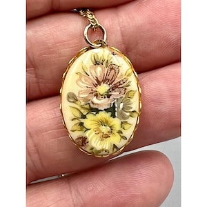 May include: A gold chain necklace with an oval pendant featuring a hand-painted floral design. The pendant is white with yellow, pink, and brown flowers.