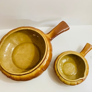 Puede incluir: Dos cuencos de cerámica marrón con asas. El cuenco más grande es ligeramente más ancho y tiene una forma más redondeada. El cuenco más pequeño es más compacto y tiene una forma más puntiaguda.