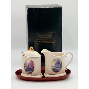 May include: A Thomas Kinkade Painter of Light sugar and creamer set featuring cream-colored ceramic pieces with gold trim and oval landscape decals. The set includes a lidded sugar bowl, a creamer pitcher, and a dark red oval tray, all in a black box.