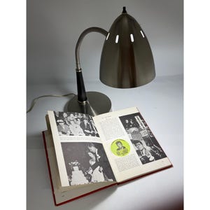 Puede incluir: Una lámpara de escritorio vintage con una pantalla cónica plateada y un cuello flexible. La lámpara está colocada sobre un libro abierto con fotografías y texto en blanco y negro. El libro tiene una cubierta roja.