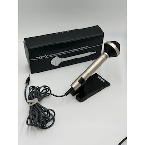 Puede incluir: Un micr&oacute;fono de condensador electret Sony ECM-198 con cuerpo met&aacute;lico plateado y cabezal negro, montado sobre un soporte negro. El micr&oacute;fono incluye un cable gris largo con conector jack y se presenta con su caja rectangular negra original.