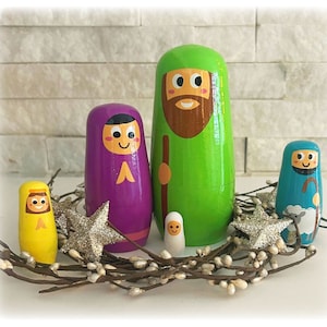 May include: A set of five wooden nesting dolls depicting the Nativity scene. The dolls are painted in bright colors, including green, purple, blue, and yellow. The largest doll is a green-clad figure with a brown beard, representing Joseph. The next largest doll is a purple-clad figure with a pink face, representing Mary. The smallest doll is a white-clad figure with a yellow halo, representing baby Jesus. The other two dolls are a blue-clad figure with a brown beard, representing a shepherd, and a yellow-clad figure with a brown beard, representing an angel.