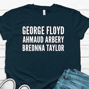 May include: A navy blue t-shirt with the names "GEORGE FLOYD, AHMAUD ARBERY, BREONNA TAYLOR" in white block letters. The shirt is paired with blue jeans and white canvas sneakers.